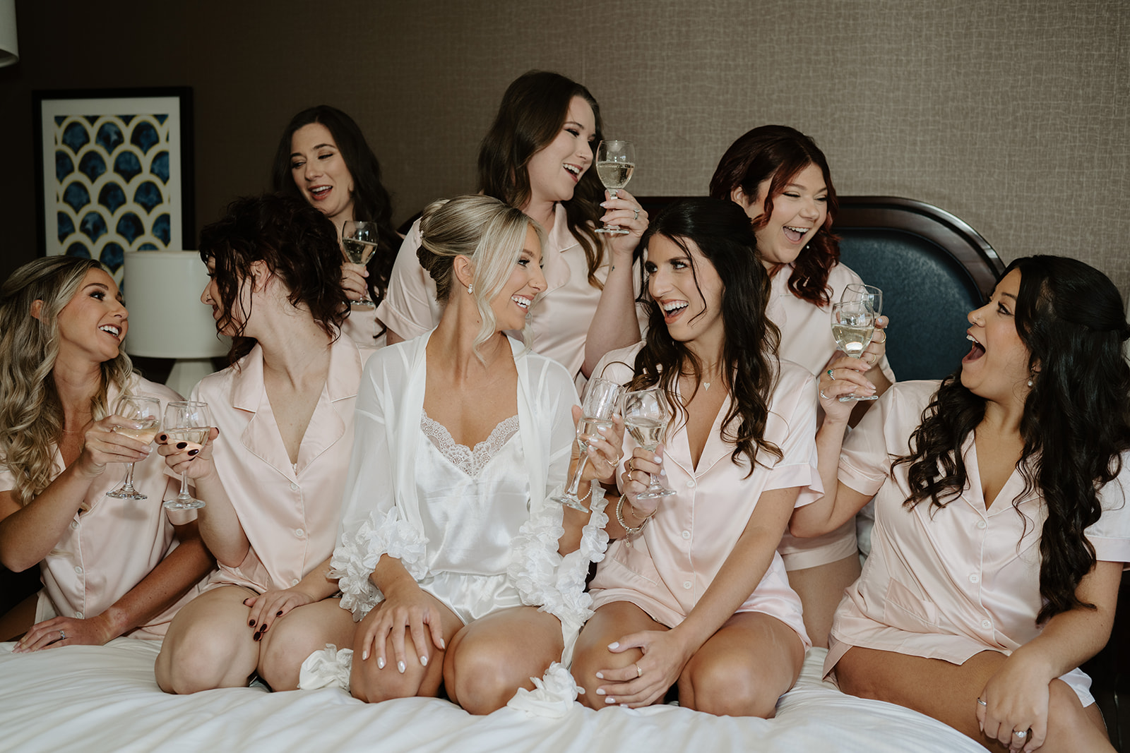 Bride and bridesmaids in pink satin pajamas toast champagne on a bed, sharing laughs during wedding prep.